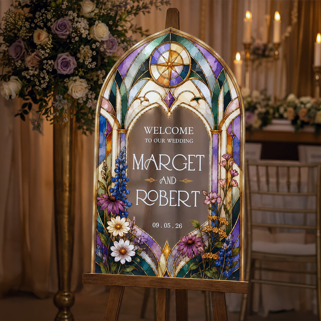 A stained-glass-style wedding sign on a wooden easel in a warmly lit reception venue. The sign features an arched Gothic window design with purple, blue, green, and gold accents, floral illustrations, and customizable text. Victorian Wildflower Stained Glass Wedding Welcome Sign, Frosted Acrylic