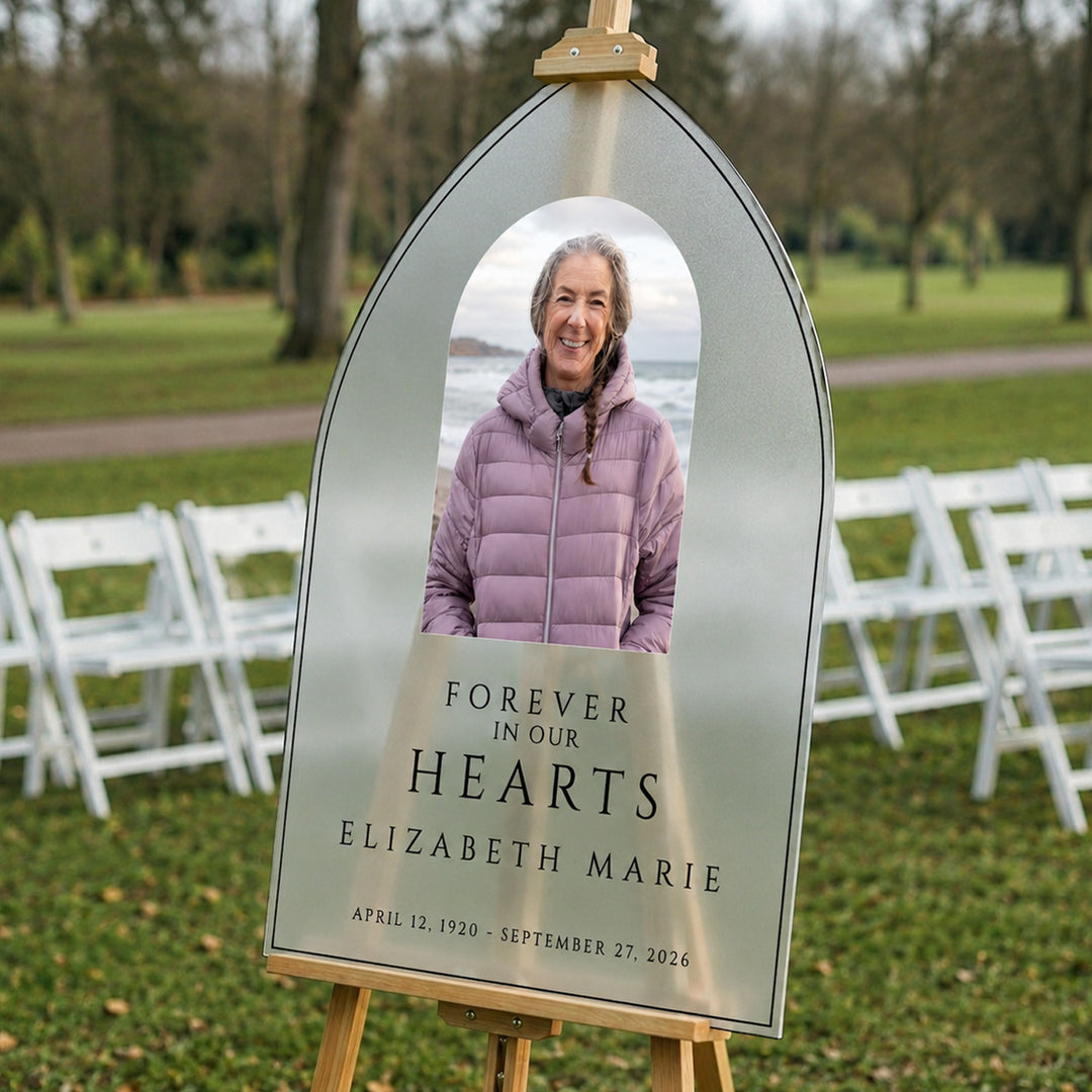 A memorial sign on an easel in a park, featuring a photo of a smiling woman in a purple puffer jacket, with the text 'FOREVER IN OUR HEARTS' and memorial dates below.