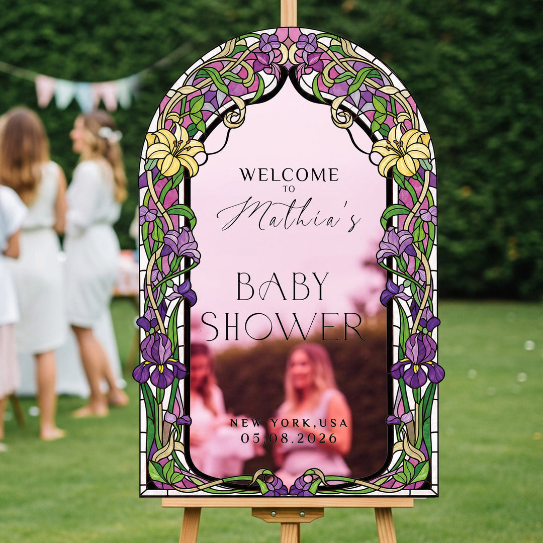 A decorative arched welcome sign for a baby shower displayed on a wooden easel outdoors. The sign has a stained-glass-style border with purple flowers, green leaves, and yellow accents, with custom text. Pink mirror acrylic.