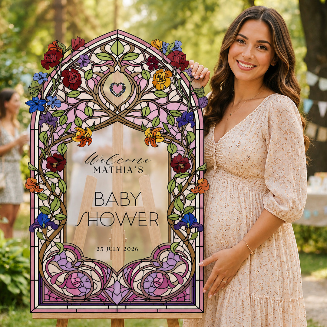 A woman in a light beige dress stands outdoors holding a large stained-glass-style arch sign with custom text. The sign features floral and scrollwork designs in pink, purple, blue, red, yellow, and green, with a heart at the top. Colorful Wildflower Stained Glass Baby Shower Welcome Sign, clear acrylic.