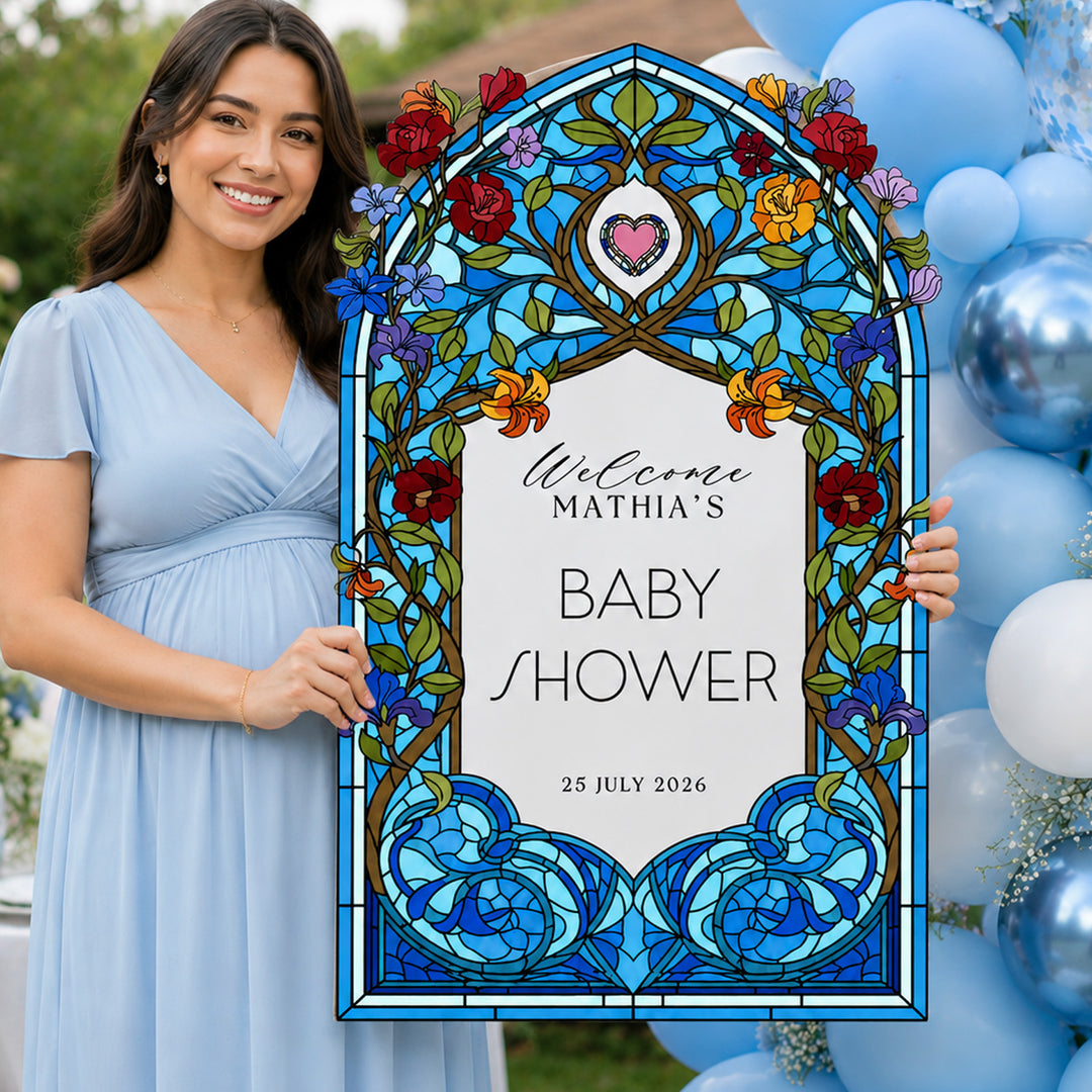 A woman in a light blue dress holds a large decorative arch sign that reads 'Welcome Mathia's Baby Shower' with a stained-glass-style blue floral design. The background shows blue and white balloons outdoors. Blue Wildflower Stained Glass Baby Shower Welcome Sign, premium PVC.