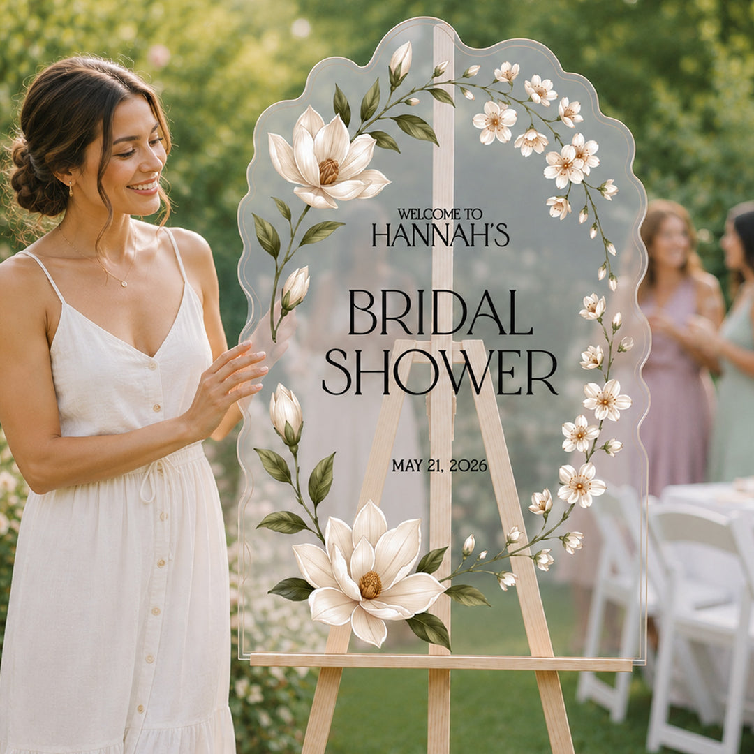 A large scalloped acrylic wedding sign on an easel in an outdoor garden setting. The sign is customizable, and is decorated with white magnolia flowers and green leaves. White Magnolia Art Nouveau Bridal Shower Welcome Sign, clear acrylic sign.