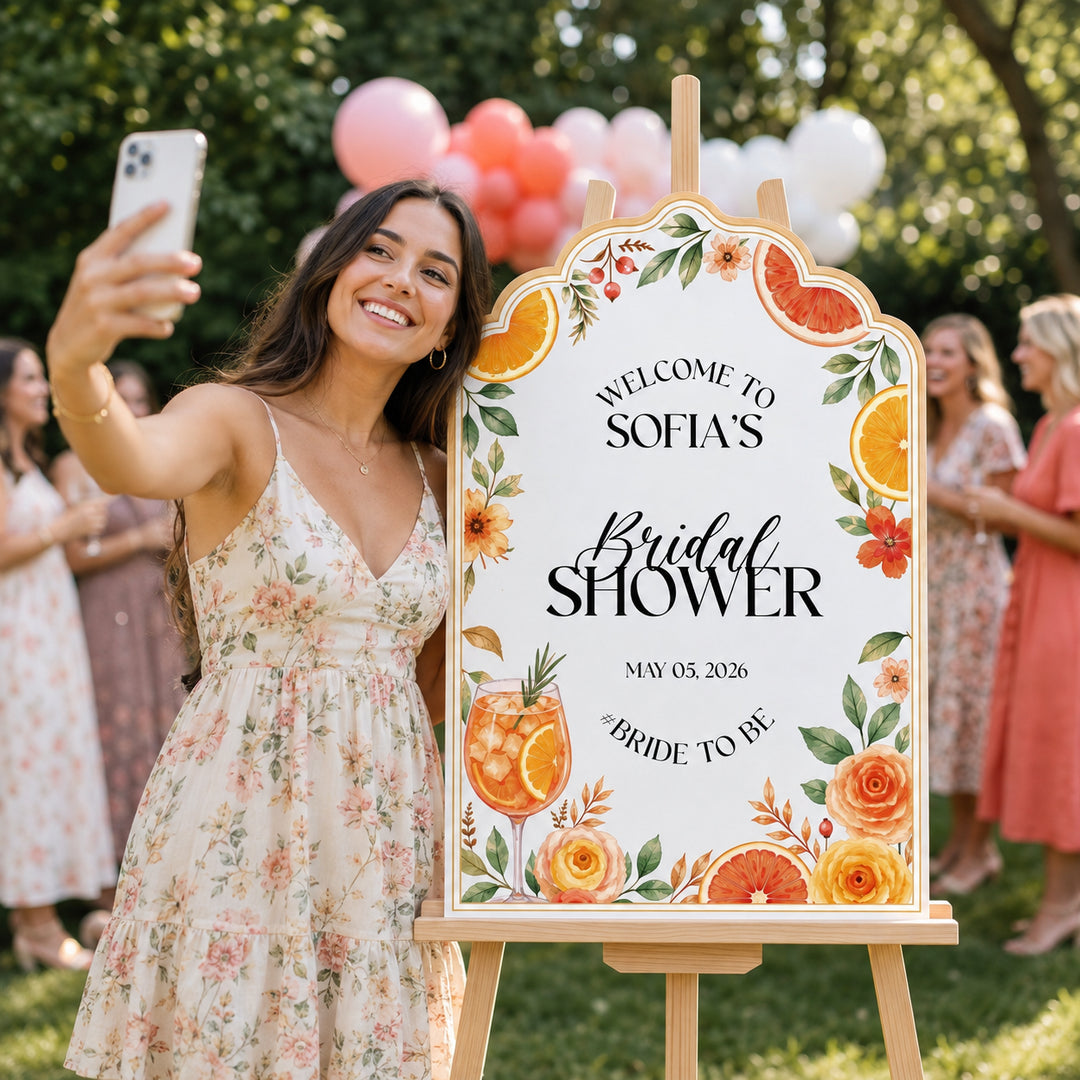A woman takes a selfie outdoors next to a decorative wooden sign on an easel that reads 'Welcome to Sofia's Bridal Shower' with floral and citrus illustrations. Guests and balloons are blurred in the background.