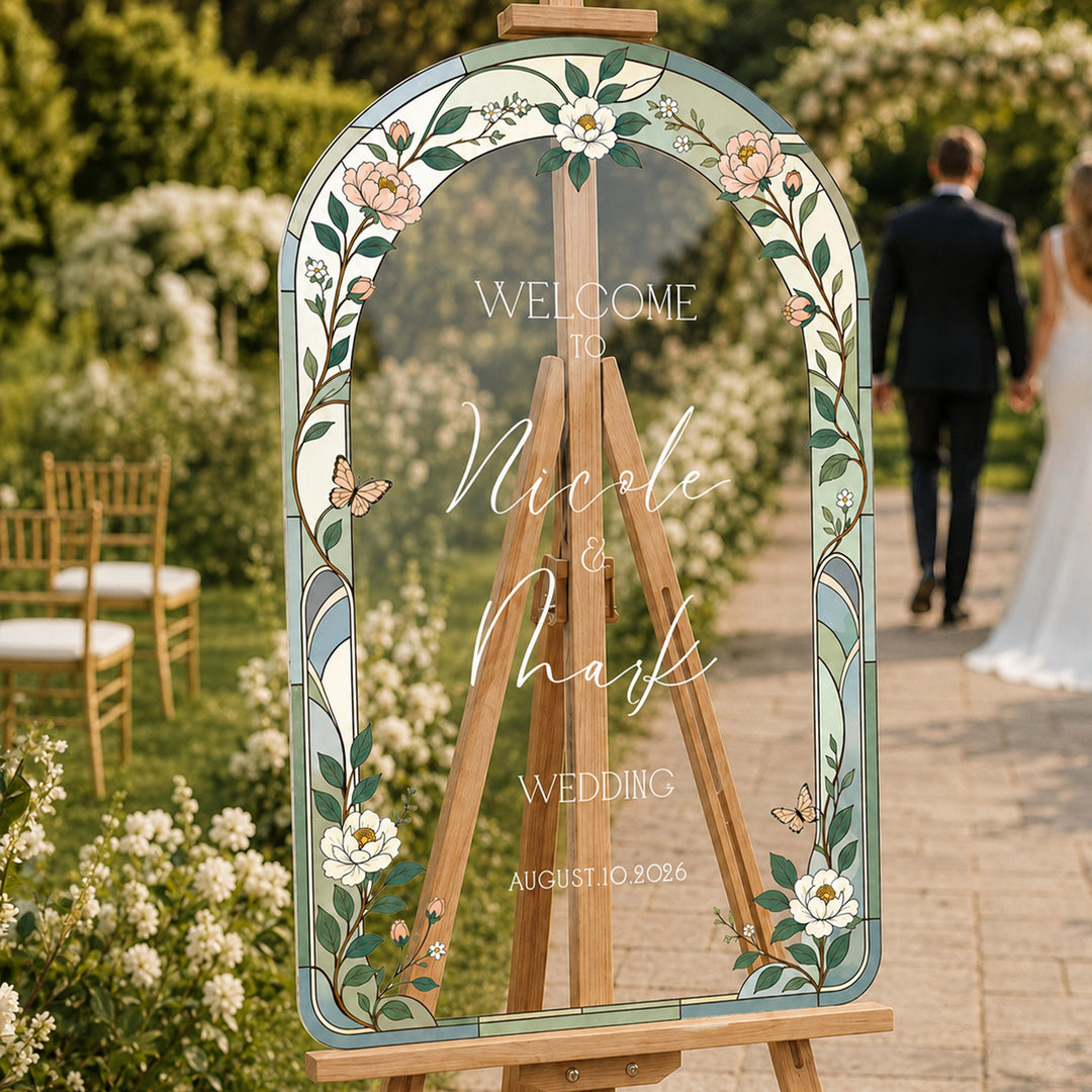 Sage wildflower stained glass wedding welcome sign on easel at garden ceremony with couple.