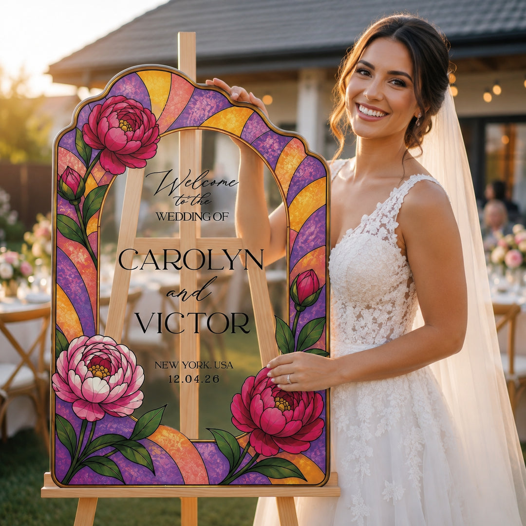 A smiling bride in a white lace wedding dress stands beside a large decorative wedding sign on an easel. The sign has an arched top and colorful stained-glass-style border in purple, yellow, orange, and pink, with floral illustrations and the text 'Welcome to the wedding of Carolyn and Victor' and the date '12.